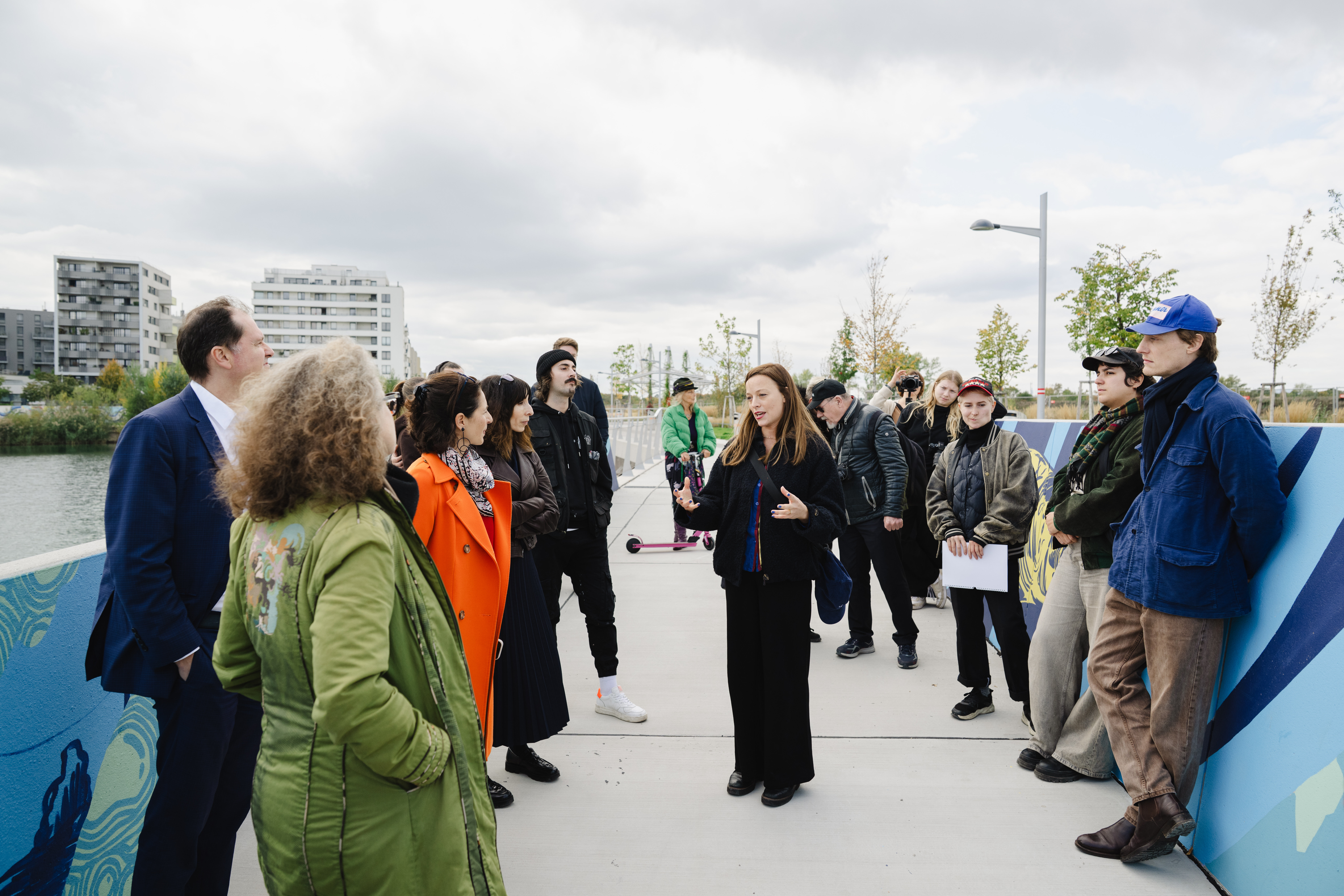 Menschen stehen auf einer Brücke mit bunten Wandmalereien und hören einer Frau zu, die spricht. Im Hintergrund sind moderne Wohngebäude zu sehen.