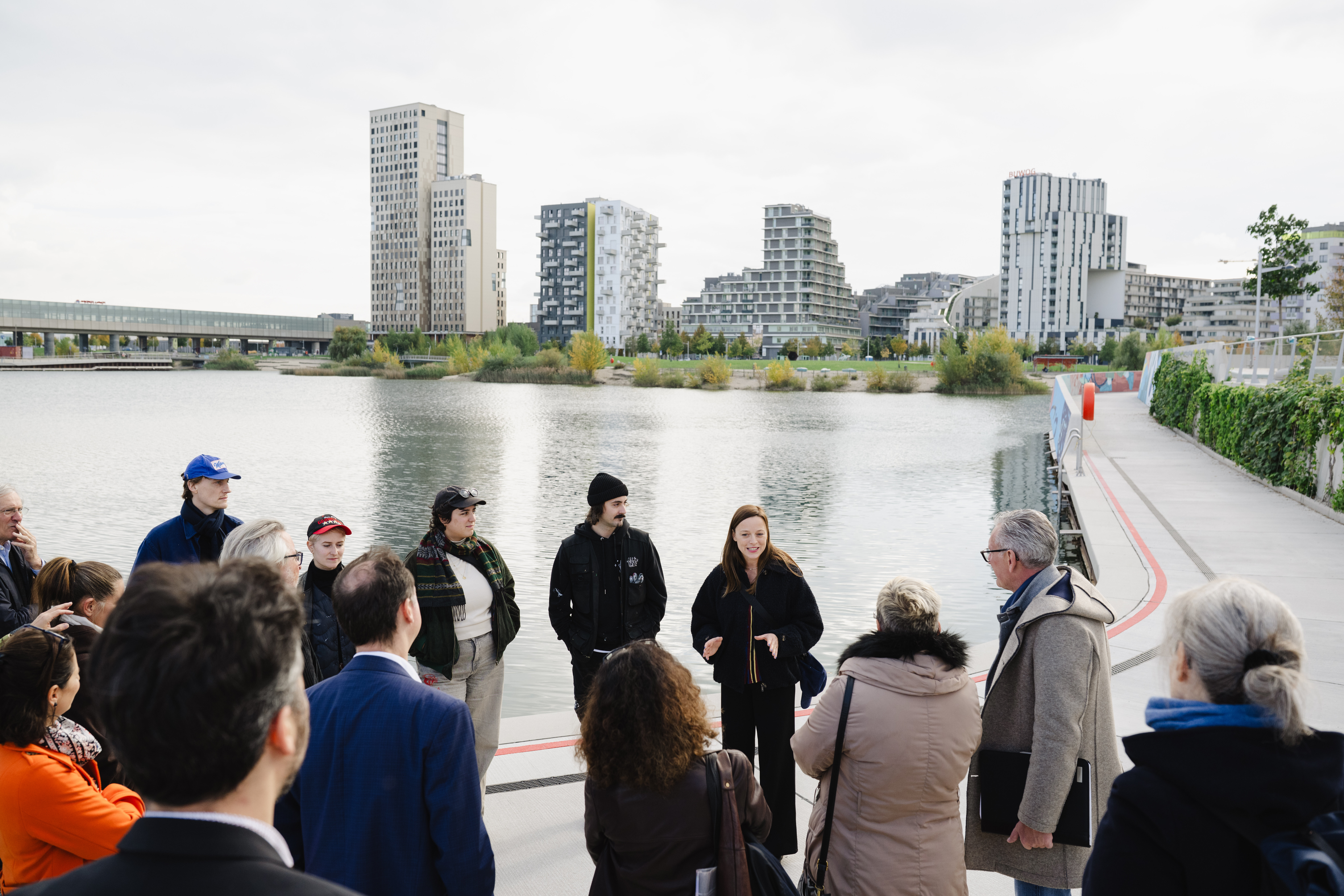 Eine Gruppe von Menschen steht am Ufer eines Sees mit moderner Stadtarchitektur im Hintergrund. Eine Frau spricht zur Gruppe, die ihr aufmerksam zuhört.