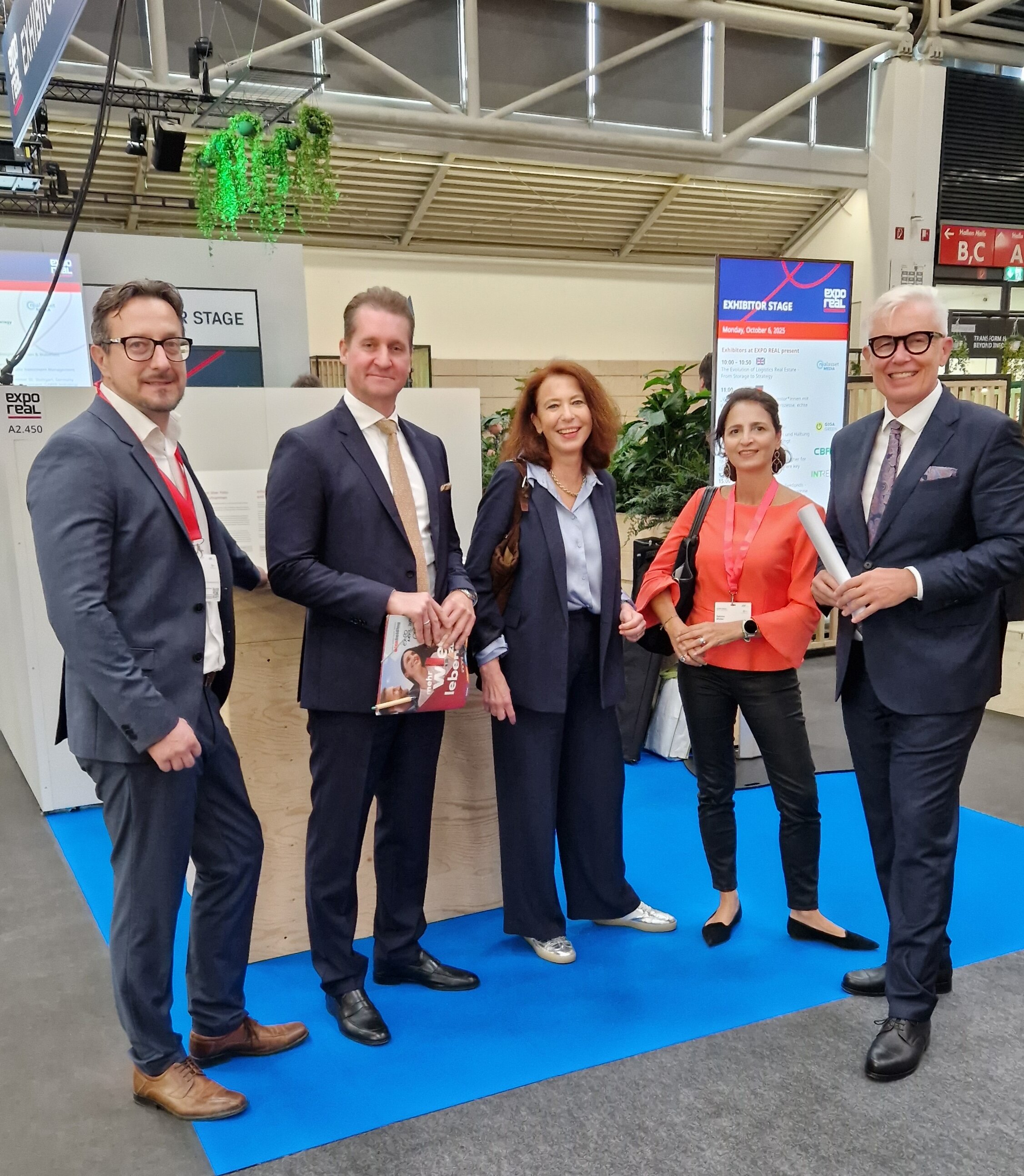 Gruppenfoto: Drei Männer im Anzug und zwei Frauen im Kostüm stehen vor einem Messestand und lächeln in die Kamera