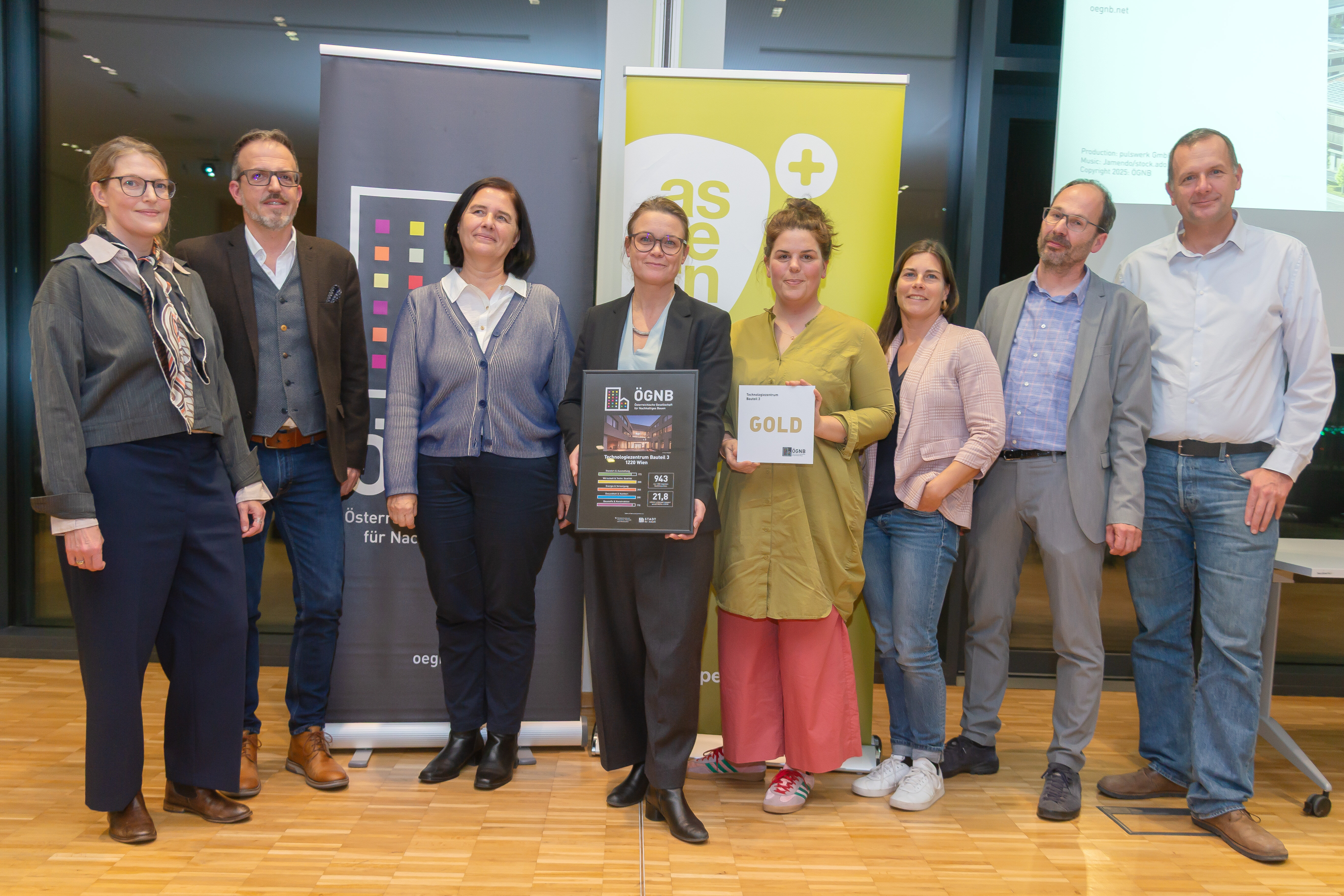 Fünf Frauen und 3 Männer stehen nebeneinander und blicken in die Kamera, die beiden Frauen in der Mitte halten einen Bilderrahmen, eine mit einem Zertifikat mit der Aufschrift ÖGNB, eine mit einem Schild mit der Aufschrift GOLD