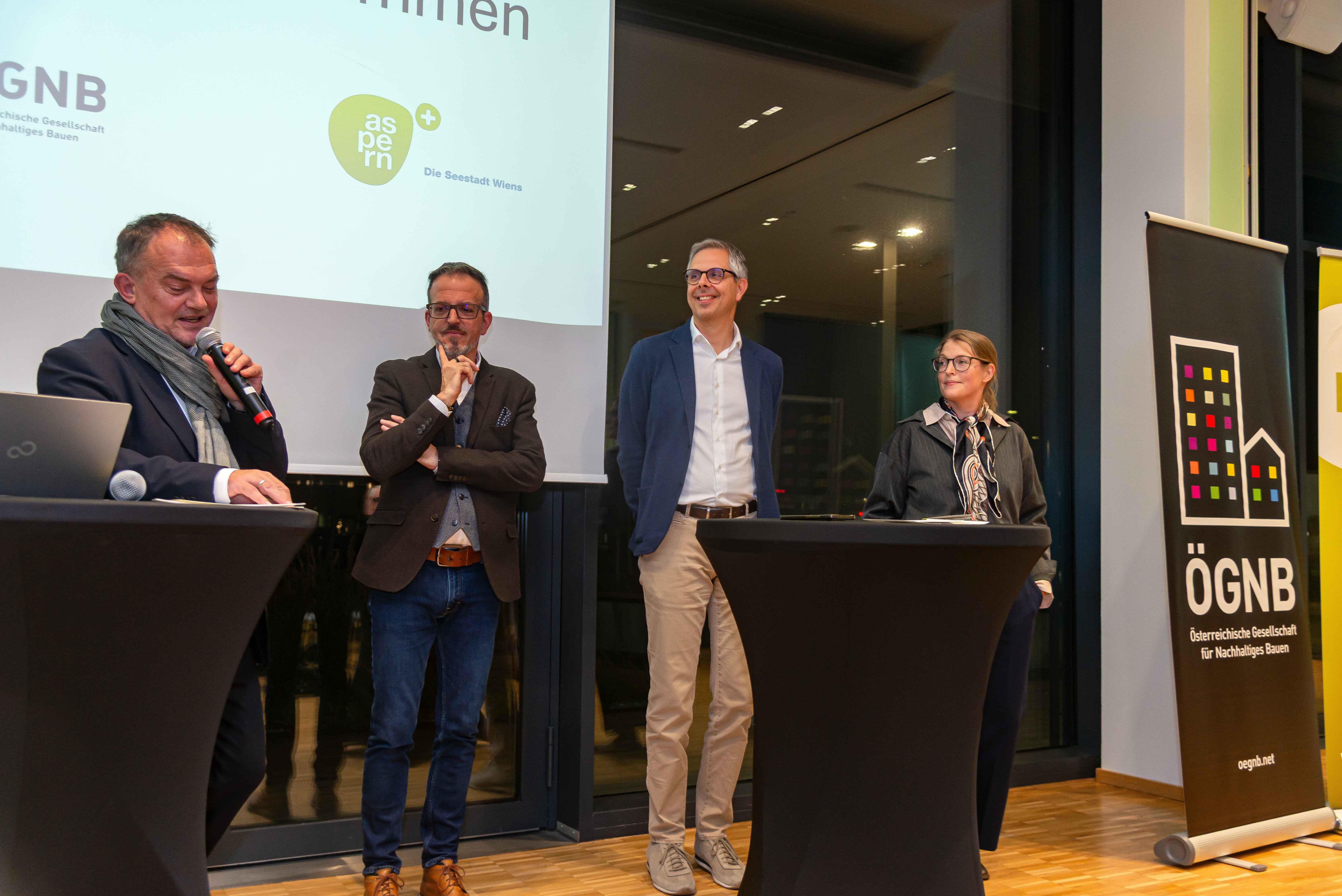 Vier Männer stehen auf dem Podium, der Mann ganz links hat ein Mikrofon in der Hand und spricht, im linken Teil des Bildes sieht man eine Leinwand und das Wien3420-Logo