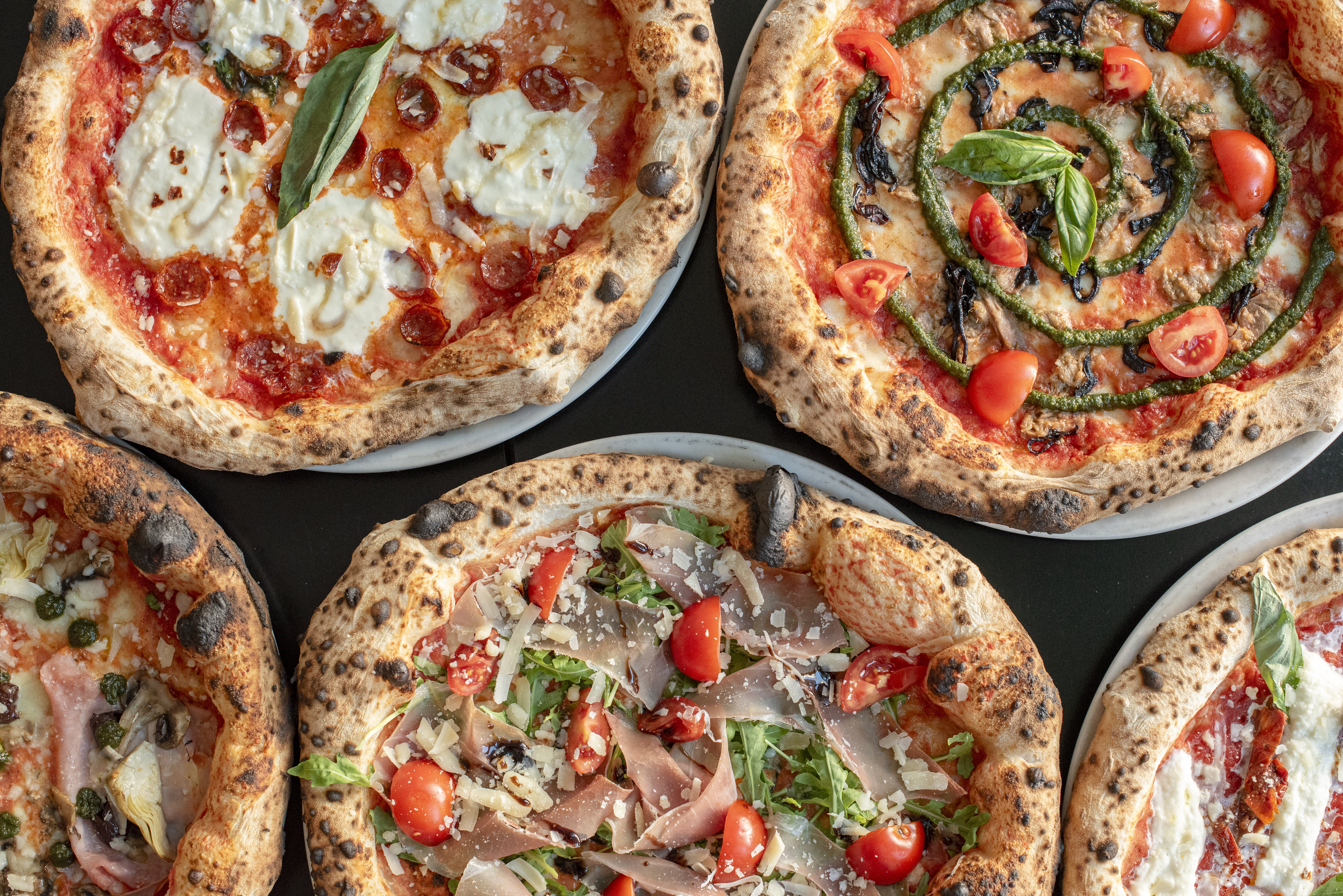 Mehrere neapolitanische Pizzen mit verschiedenen Belägen – darunter Tomaten, Burrata, Pesto, Gemüse und Schinken – liegen dicht nebeneinander und sind aus der Vogelperspektive fotografiert.