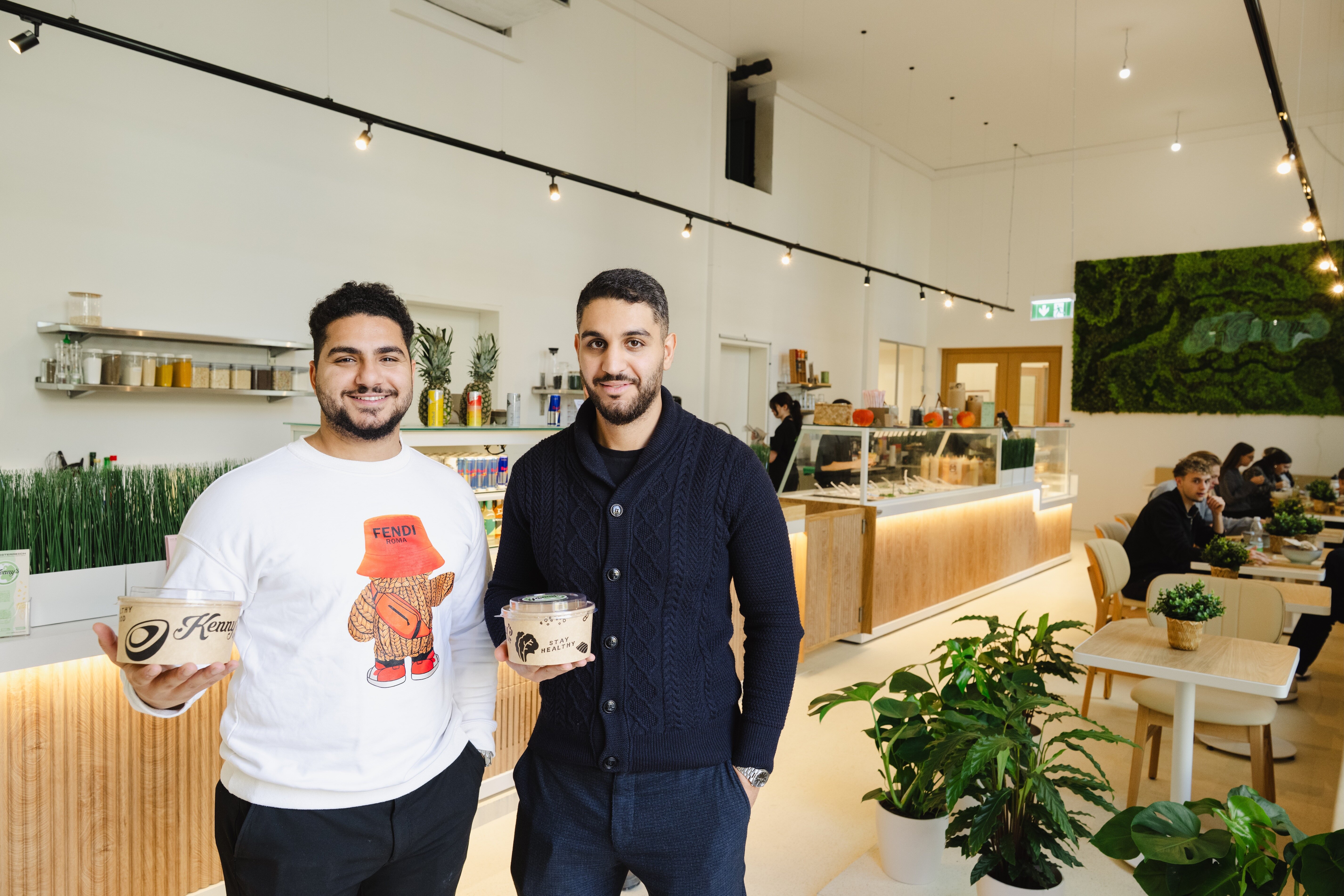 Zwei junge Männer stehen lächelnd in einem modernen, hellen Restaurant und halten Take-away-Bowls in den Händen. Im Hintergrund ist eine lange Theke mit frischen Zutaten sowie Gäste, die an Tischen sitzen.
