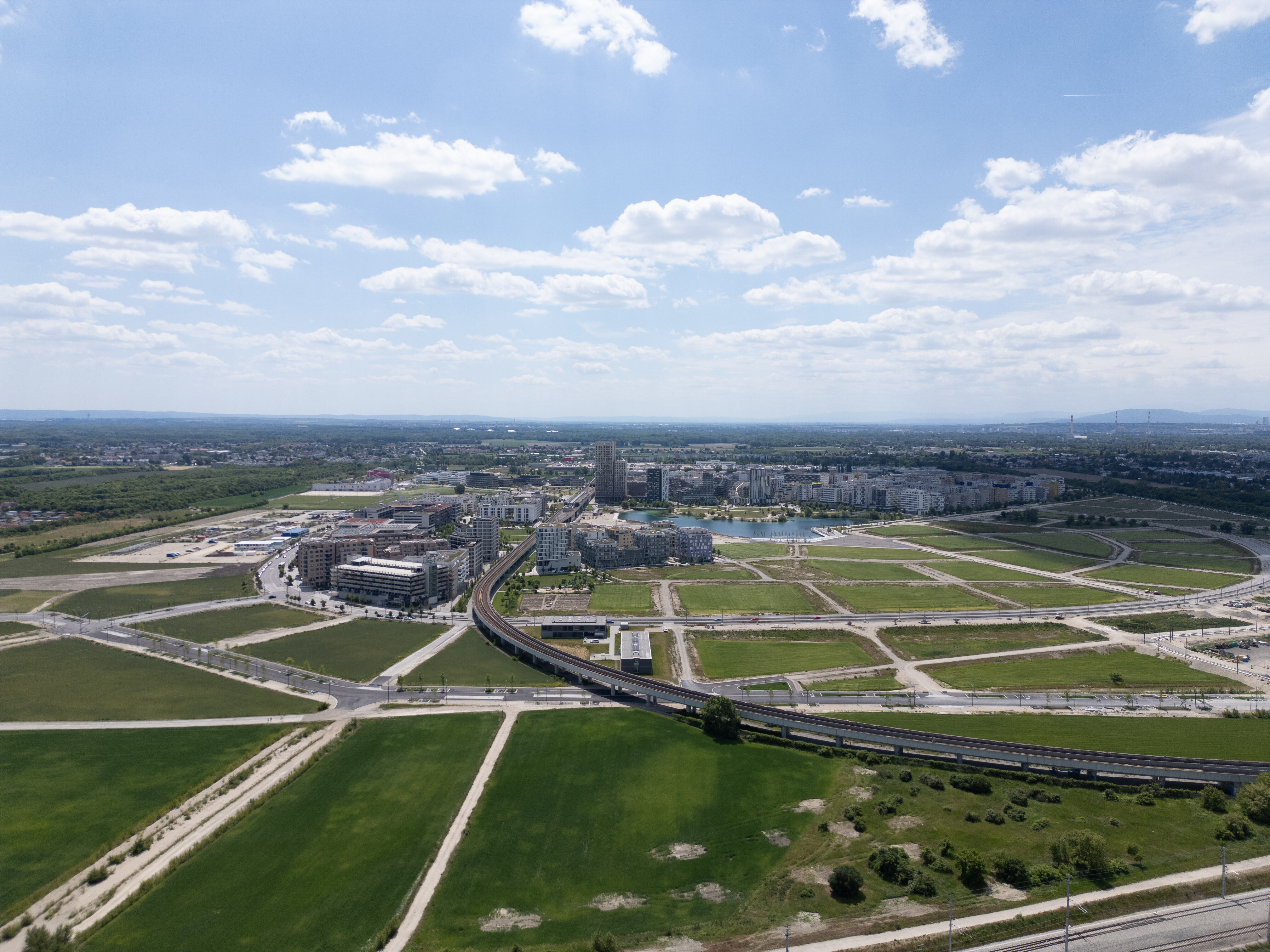 Luftaufnahme der aspern Seestadt mit Wohngebäuden, Grünflächen, Straßen und U-Bahn-Trasse unter blauem Himmel.