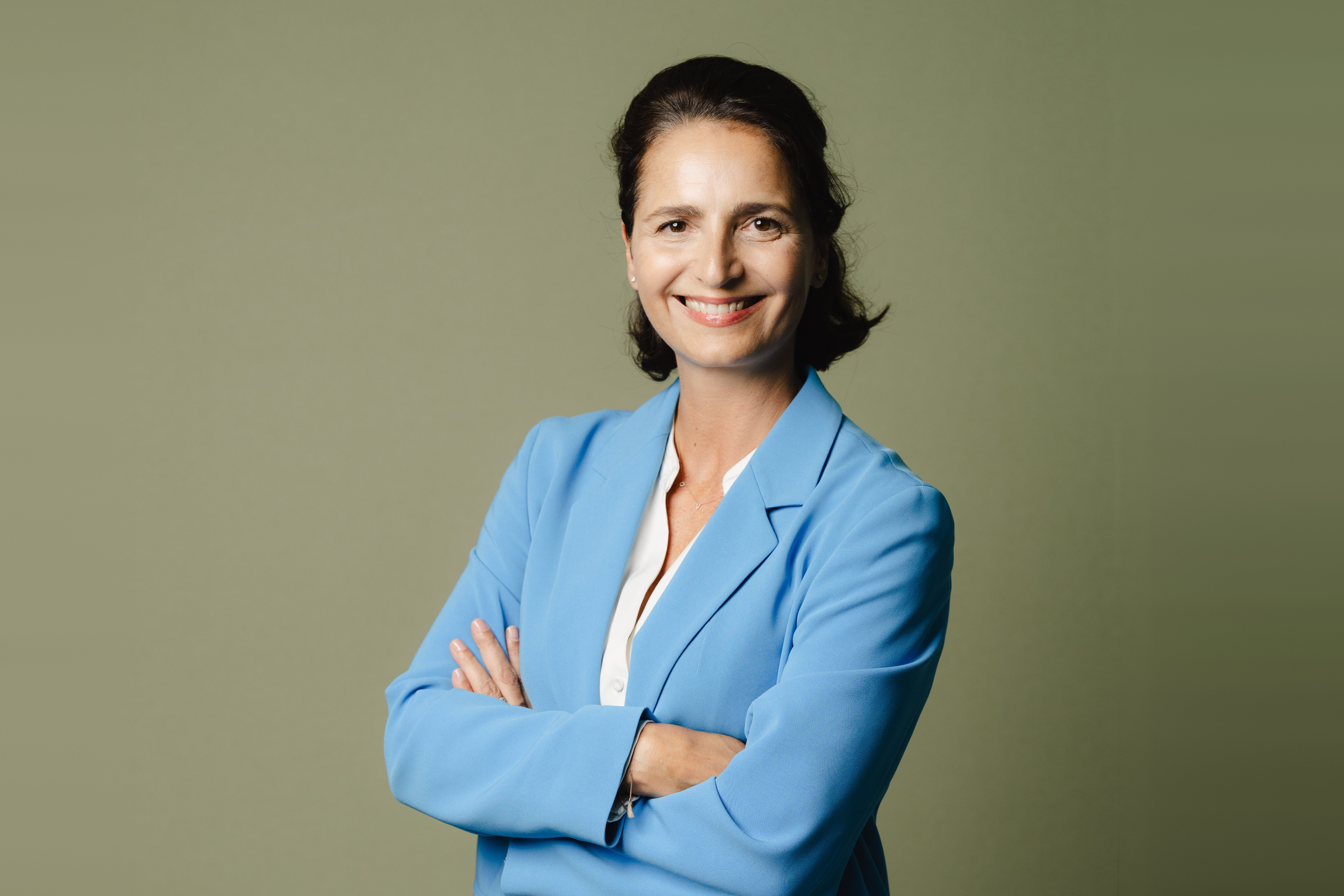 Porträt von Sabine Müller in blauem Blazer vor olivgrünem Hintergrund, lächelnd mit verschränkten Armen.